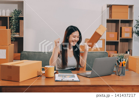 Startup SME small business entrepreneur of freelance Asian woman wearing apron using laptop and box to receive and review orders online to prepare to pack sell to customers, online sme business ideas. Startup SME small business entrepreneur of freelance Asian woman wearing apron using laptop and box to receive and review orders online to prepare to pack sell to customers, online sme business ideas. 141437217