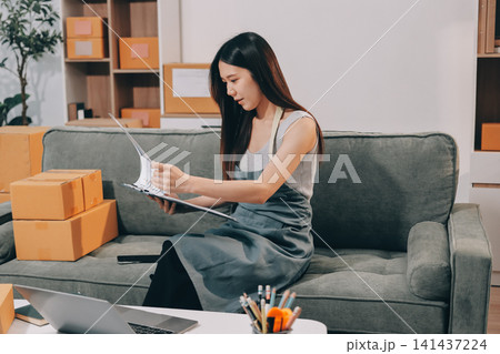 Startup SME small business entrepreneur of freelance Asian woman wearing apron using laptop and box to receive and review orders online to prepare to pack sell to customers, online sme business ideas. Startup SME small business entrepreneur of freelance Asian woman wearing apron using laptop and box to receive and review orders online to prepare to pack sell to customers, online sme business ideas. 141437224