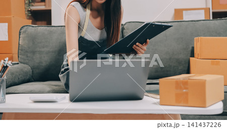 Startup SME small business entrepreneur of freelance Asian woman wearing apron using laptop and box to receive and review orders online to prepare to pack sell to customers, online sme business ideas. Startup SME small business entrepreneur of freelance Asian woman wearing apron using laptop and box to receive and review orders online to prepare to pack sell to customers, online sme business ideas. 141437226