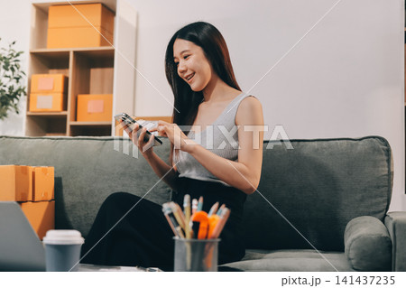 Startup SME small business entrepreneur of freelance Asian woman wearing apron using laptop and box to receive and review orders online to prepare to pack sell to customers, online sme business ideas. Startup SME small business entrepreneur of freelance Asian woman wearing apron using laptop and box to receive and review orders online to prepare to pack sell to customers, online sme business ideas. 141437235