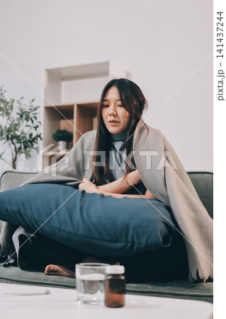 sick asian young woman sneezing into tissue paper while covered with a blanket and sitting on sofa in the living room sick asian young woman sneezing into tissue paper while covered with a blanket and sitting on sofa in the living room 141437244