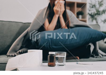 sick asian young woman sneezing into tissue paper while covered with a blanket and sitting on sofa in the living room sick asian young woman sneezing into tissue paper while covered with a blanket and sitting on sofa in the living room 141437245