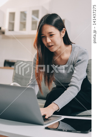Freelance Asian woman holding mobile phone typing on laptop and working from home. Happy young girl sitting on sofa in living room. Freelance Asian woman holding mobile phone typing on laptop and working from home. Happy young girl sitting on sofa in living room. 141437275
