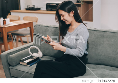 Freelance Asian woman holding mobile phone typing on laptop and working from home. Happy young girl sitting on sofa in living room. Freelance Asian woman holding mobile phone typing on laptop and working from home. Happy young girl sitting on sofa in living room. 141437276