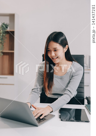 Young asian businesswoman is smiling and working on a laptop while sitting at her desk in a modern office Young asian businesswoman is smiling and working on a laptop while sitting at her desk in a modern office 141437281