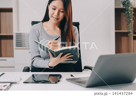 Young asian businesswoman is smiling and working on a laptop while sitting at her desk in a modern office Young asian businesswoman is smiling and working on a laptop while sitting at her desk in a modern office 141437284