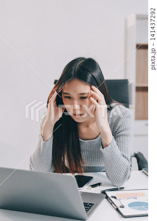 Young Asian woman sits at office desk rubbings her eyes and temples feeling pain. Stress. And exhaustion from long hours working on the computer Young Asian woman sits at office desk rubbings her eyes and temples feeling pain. Stress. And exhaustion from long hours working on the computer 141437292