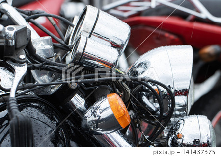 Detailed close-up view of shiny chrome components of modern motorcycle, featuring handlebars, instrument gauges, and bright headlights covered with numerous water droplets, reflecting wet weather 141437375