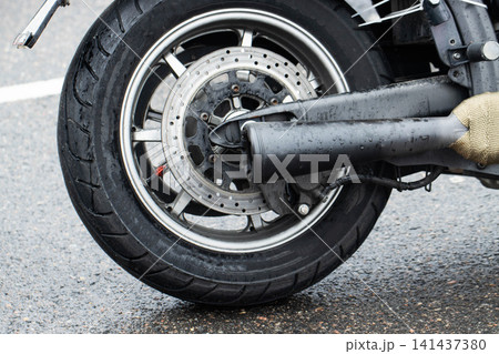 Rear wheel of motorcycle on wet asphalt road surface, detail of tire tread, brake disc, and exhaust pipe with raindrops, vehicle parked outdoors after rain Rear wheel of motorcycle on wet asphalt road surface, detail of tire tread, brake disc, and exhaust pipe with raindrops, vehicle parked outdoors after rain 141437380