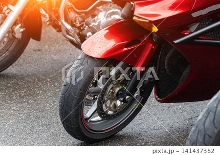 Close up view of front wheel, tire, and red fender of sport motorcycle with water drops on wet asphalt ground, blurred other motorcycles in background with warm light lens flare. Close up view of front wheel, tire, and red fender of sport motorcycle with water drops on wet asphalt ground, blurred other motorcycles in background with warm light lens flare. 141437382