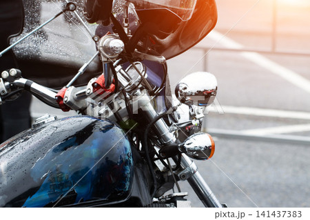 Close-up detail of motorcycle front section with handlebars, fuel tank, chrome elements, and rearview mirror showing raindrops, sun flare on windshield, custom paint artwork on tank, street background 141437383