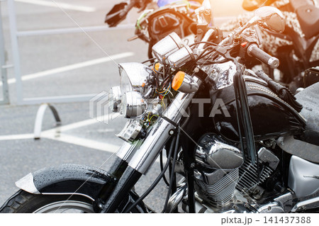 Black cruiser motorcycle with chrome details parked outdoors, covered in raindrops after rain, reflecting ambient light on wet surface, showcasing vehicle front section with headlights and handlebars. 141437388