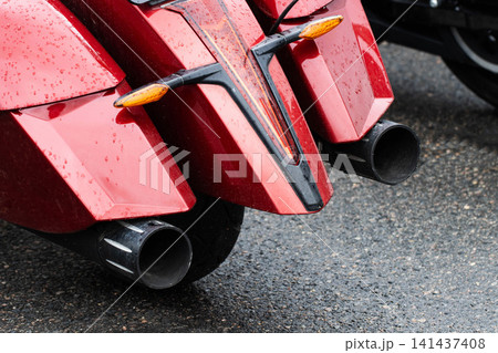 Rear view of shiny red motorcycle fender with visible raindrops, featuring dual chrome exhaust pipes and orange turn signals on wet asphalt surface, presenting aftermath of rain. Rear view of shiny red motorcycle fender with visible raindrops, featuring dual chrome exhaust pipes and orange turn signals on wet asphalt surface, presenting aftermath of rain. 141437408