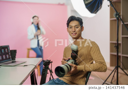 Smiling young Asian male photographer holding professional camera in studio Smiling young Asian male photographer holding professional camera in studio 141437546