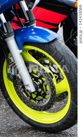 Close-up of front wheel of motorcycle with vibrant neon yellow rim on wet asphalt after rain. Blue fender, silver forks, red body panel visible, showcasing detail of braking system and tire tread. 141438066