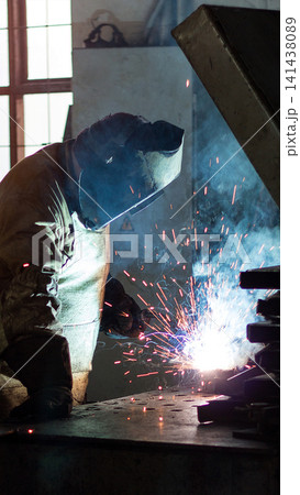 Skilled worker performing arc welding process in industrial workshop creating bright sparks and smoke, wearing protective helmet and gloves for safety, close up view vertical photo Skilled worker performing arc welding process in industrial workshop creating bright sparks and smoke, wearing protective helmet and gloves for safety, close up view vertical photo 141438089