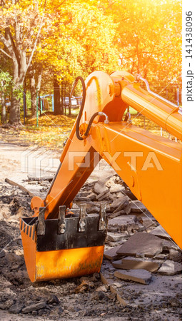 Close up view of orange excavator arm and bucket on broken asphalt ground, autumn leaves and golden sunlight in background, construction equipment work site vertical photo 141438096