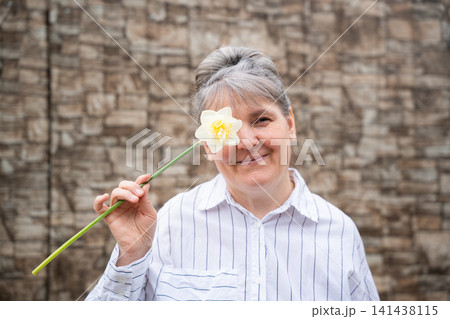 Smiling mature woman holding a daffodil against a rustic stone wall. Positive aging mindfulness and women's health awareness Smiling mature woman holding a daffodil against a rustic stone wall. Positive aging mindfulness and women's health awareness 141438115