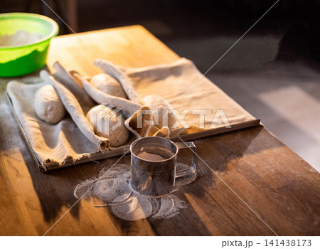 Freshly kneaded bread dough rounds resting on linen cloth in proofing basket, preparation for baking on wooden kitchen table with flour dusting and sifter, homemade bakery goods production Freshly kneaded bread dough rounds resting on linen cloth in proofing basket, preparation for baking on wooden kitchen table with flour dusting and sifter, homemade bakery goods production 141438173
