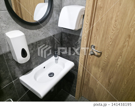 Modern public restroom interior showing white ceramic sink, chrome faucet, wall mounted soap dispenser, hand dryer, round mirror, dark gray tiled walls and wooden door Modern public restroom interior showing white ceramic sink, chrome faucet, wall mounted soap dispenser, hand dryer, round mirror, dark gray tiled walls and wooden door 141438195
