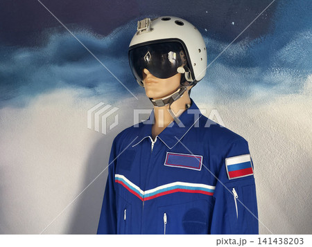 Mannequin display of pilot flight suit and helmet with visor, featuring blue jumpsuit, white helmet, and chest stripes against painted wall background, showcasing aviation attire. Mannequin display of pilot flight suit and helmet with visor, featuring blue jumpsuit, white helmet, and chest stripes against painted wall background, showcasing aviation attire. 141438203