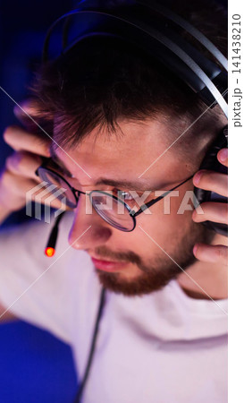 Young man in glasses and headphones with illuminated microphone adjusts headset, concentrated expression during online gaming or communication session, blue background light, vertical photo Young man in glasses and headphones with illuminated microphone adjusts headset, concentrated expression during online gaming or communication session, blue background light, vertical photo 141438210