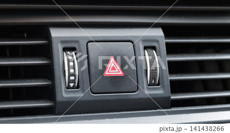 Vehicle dashboard close-up showing hazard warning light button with red triangle symbol, surrounded by car interior air conditioning vents and rotary controls for safety feature Vehicle dashboard close-up showing hazard warning light button with red triangle symbol, surrounded by car interior air conditioning vents and rotary controls for safety feature 141438266