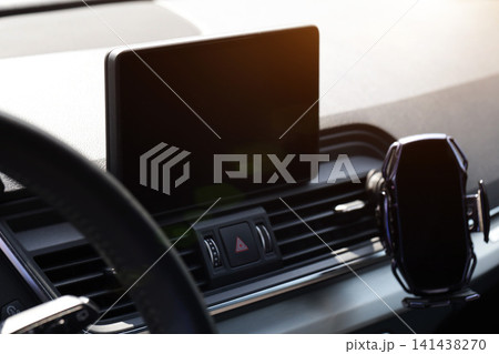 Modern car dashboard detail showing black infotainment screen, hazard warning button, and smartphone mount with air conditioning vents, sunlight flare creating reflection. Modern car dashboard detail showing black infotainment screen, hazard warning button, and smartphone mount with air conditioning vents, sunlight flare creating reflection. 141438270
