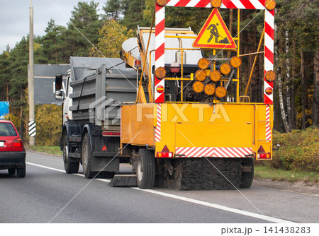 Road sweeper truck with warning sign and hazard stripes cleaning edge of asphalt road with forest background 141438283