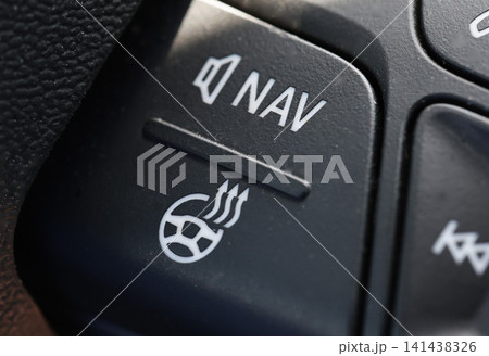 Close-up image of car steering wheel control panel button showing white icons for volume, navigation, and heated steering wheel, detail of automotive interior design 141438326