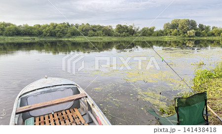 A fishing spot on a quiet river: a boat near the shore, a fishing rod, and a folding chair in front of a green forest. A fishing spot on a quiet river: a boat near the shore, a fishing rod, and a folding chair in front of a green forest. 141438418