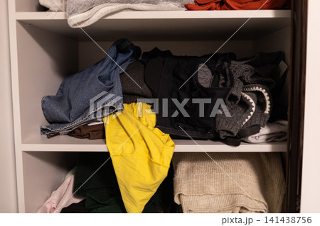 Open wardrobe shelves with casually stacked clothes, bags and a bright yellow garment, showing cluttered everyday storage and realistic home organization in a domestic interior 141438756