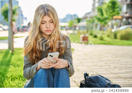 Serious teenage girl with long blonde hair sitting outdoors using smartphone 141438990