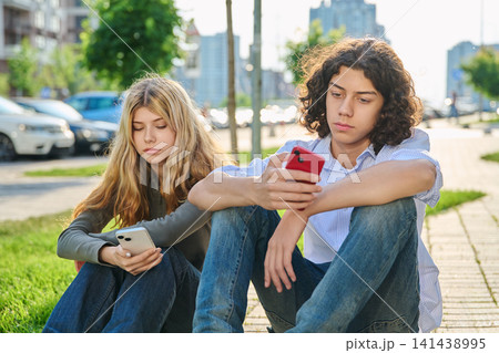 Teenage boy and girl sitting outside glued to smartphones Teenage boy and girl sitting outside glued to smartphones 141438995