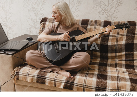 Mature woman practicing guitar beside a laptop at home Mature woman practicing guitar beside a laptop at home 141439378