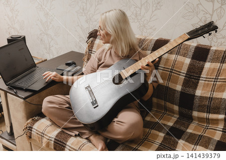 Mature woman using a laptop while holding a guitar at home Mature woman using a laptop while holding a guitar at home 141439379