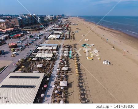 Beautiful high altitude aerial panoramic perspective of beach resort clubs with straw parasols giant sunglasses sculpture and promenade along a sandy coast under a blue sky Beautiful high altitude aerial panoramic perspective of beach resort clubs with straw parasols giant sunglasses sculpture and promenade along a sandy coast under a blue sky 141439830