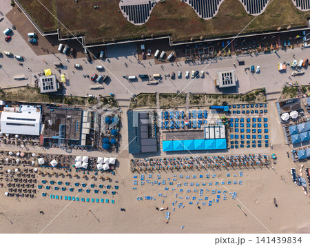 Beautiful top down high altitude aerial panoramic perspective of beach resort clubs with blue sunbeds umbrellas green roofs and parked vehicles along a seaside promenade under bright sunlight Beautiful top down high altitude aerial panoramic perspective of beach resort clubs with blue sunbeds umbrellas green roofs and parked vehicles along a seaside promenade under bright sunlight 141439834