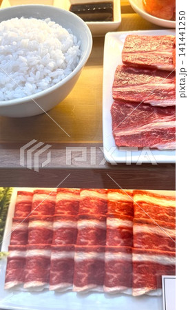 Steamed rice bowl beside sliced raw beef on white plates with warm lighting, showing Japanese barbecue meal preparation on wooden counter with glossy texture and fresh color Steamed rice bowl beside sliced raw beef on white plates with warm lighting, showing Japanese barbecue meal preparation on wooden counter with glossy texture and fresh color 141441250