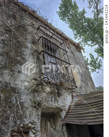 A damaged window shows protective iron bars with a wooden shutter above a rusty roof on a neglected building. Plants grow around the structure, hinting at years of abandonment. 141442114