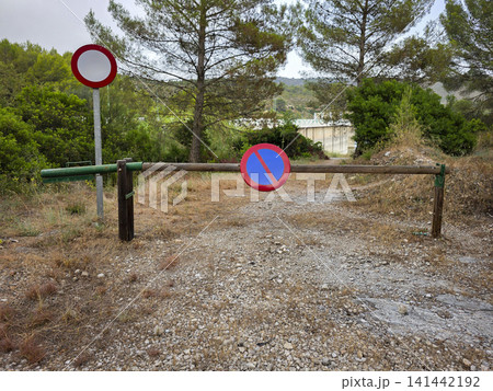 A wooden barrier gate is closed across a gravel path leading to a dam. No parking and no entry signs are visible beside the gate. Trees and bushes surround the area. A wooden barrier gate is closed across a gravel path leading to a dam. No parking and no entry signs are visible beside the gate. Trees and bushes surround the area. 141442192