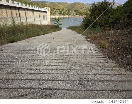 A concrete slipway with grooves leads down to the water at Beniarres Reservoir. The large dam structure is seen beside the water. Green vegetation grows along the path. A concrete slipway with grooves leads down to the water at Beniarres Reservoir. The large dam structure is seen beside the water. Green vegetation grows along the path. 141442194