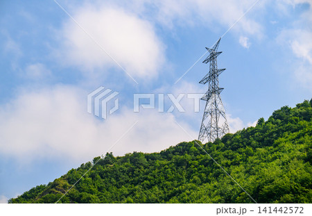 Power line tower rises above forested mountain peak beneath partly cloudy sky Power line tower rises above forested mountain peak beneath partly cloudy sky 141442572