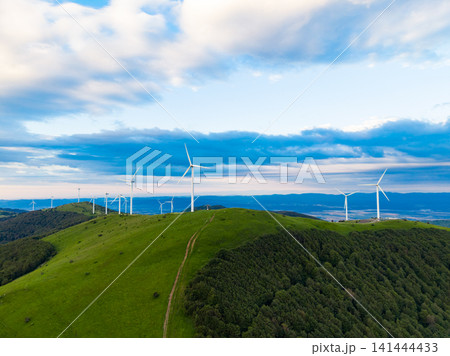 Wind turbines stand in rows on a green hill under a blue sky with fluffy clouds in the background during daylight hours 141444433