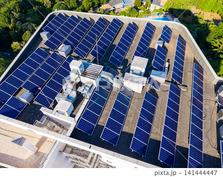 Solar panels cover a large roof in a green area with trees and a blue pool visible in the background during sunny weather Solar panels cover a large roof in a green area with trees and a blue pool visible in the background during sunny weather 141444447