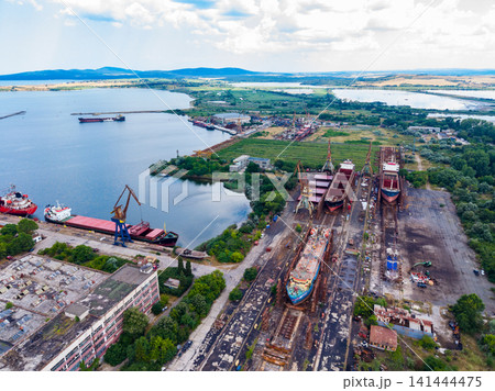 Shipping yard shows boats and cranes near water in a busy industrial area during the day with clouds in the sky Shipping yard shows boats and cranes near water in a busy industrial area during the day with clouds in the sky 141444475