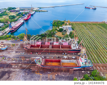 Ships docked at a harbor with cranes and boats near green fields in a coastal area during daytime 141444478