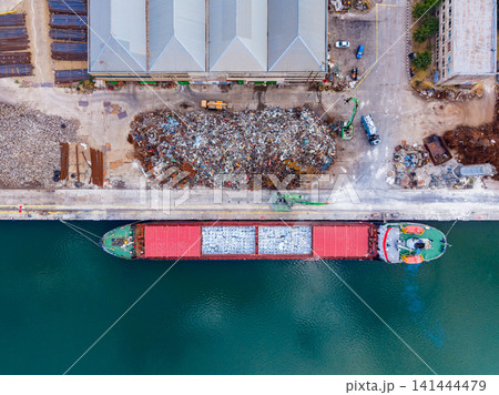 Cargo ship unloading materials at a dock near a recycling site with piles of waste and nearby buildings in an industrial area 141444479