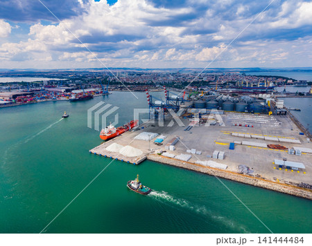 Tugboat maneuvers near busy port with cargo ships and industrial area in background Tugboat maneuvers near busy port with cargo ships and industrial area in background 141444484