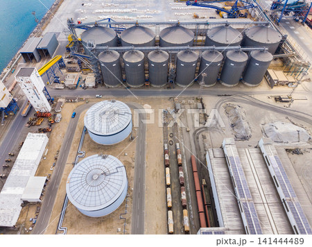 Large storage silos and tanks at an industrial site near a port with ships and machinery in the background during daytime Large storage silos and tanks at an industrial site near a port with ships and machinery in the background during daytime 141444489